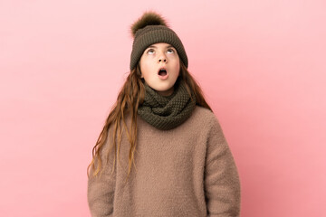 Little girl with winter hat isolated on pink background looking up and with surprised expression