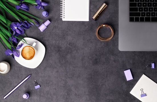 Spring women's workspace top view with beautiful iris flowers, laptop, office supplies, beauty accessorise and coffee with milk on dark grey stone background. Flat lay with copy space. Spring holidays