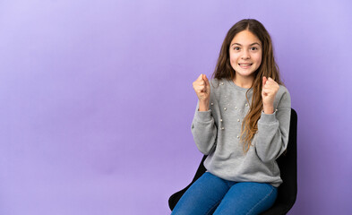 Little caucasian girl sitting on a chair isolated on purple background celebrating a victory in...