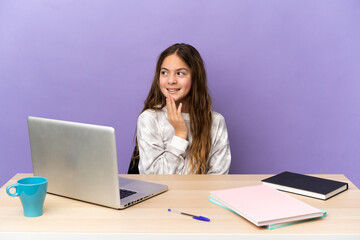 Little student girl in a workplace with a laptop isolated on purple background looking up while smiling