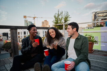 Fototapeta premium Group of multi-cultural young adults laughing at joke together. Sitting at a rooftop party in the city, good times.
