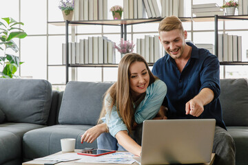 Young millennial Caucasian lover couple handsome husband and beautiful wife in casual outfit sitting together on cozy sofa watching streaming movie online from laptop notebook computer in weekend