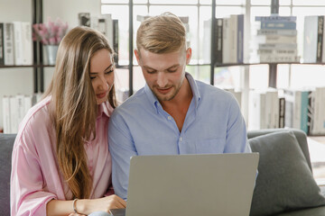 Millennial caucasian lover couple husband and wife in casual outfit sitting smiling on sofa surfing browsing internet shopping online together via laptop notebook computer in living room on weekend