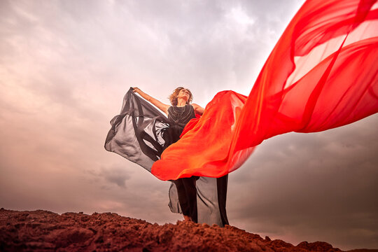 A girl in black dress with red fabric dances on sand dunes against a dramatic sky before a thunderstorm. Model posing during photo shoot on nature