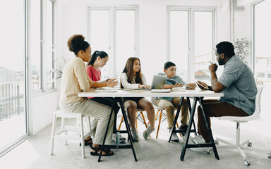 African American male teacher sitting at table with pupils teaching writing lesson in modern classroom. Intending group of elementary students sit together diligently studying and listening to tutor.