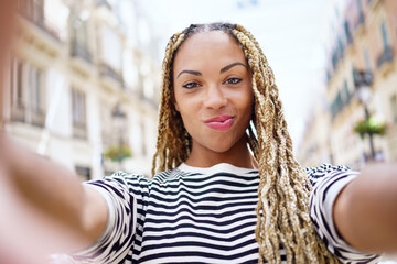 Black woman with afro braids taking a selfie in an urban street with a smartphone.