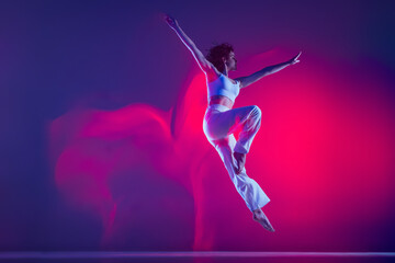 Young sportive beautiful girl, hip-hop dancer jumping isolated on purple background in pink neon light. Youth culture, style and fashion, action.