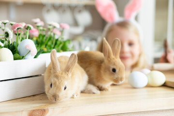 Two little Easter red rabbits sit cheerfully on the kitchen table near a cheerful little girl with ears and basket with flowers and Easter eggs. Against the backdrop of the interior of a home kitchen