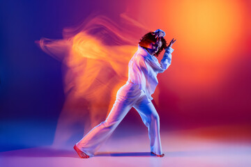Street dancing. Image of flexible young girl, hip-hop dancer in white outfit dancing hip hop isolated on blue background in yellow neon light. Youth culture, style and fashion, action.