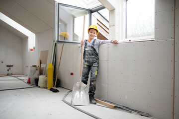 cool boy with yellow safety helmet and dungarees on construction site in a house and loft with...