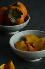 Close-up of two small ceramic blue bowls with whole and sliced persimmons on a dark background 