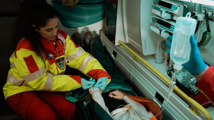 Young Ethnic Female Medic Working in an Ambulance Car, Saving a Teenagers Life, Who is In a Stretcher As They Put an IV Drip In Her Vein To Keep Her Alive on the Way To The Hospital.
