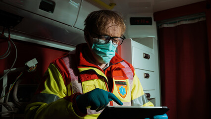 Medical Emergency Care Worker Wearing a Face Mask, Using a Tablet To Write Down Information About a Patient, Sitting At The Back Of an Ambulance Car. Hardworking Paramedic During Night Shift.