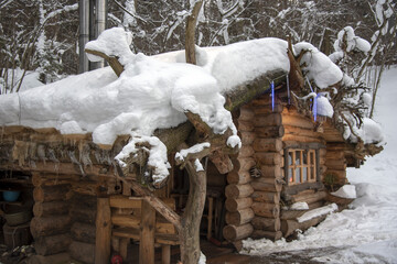 A log bathhouse, covered with snow, stands among the trees on a sunny winter day.