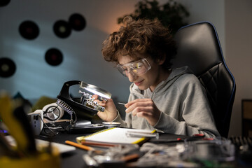 A boy is sitting at a desk in front of a soldering iron. He is repairing computer hardware engineering, technology, science, concept. A child is soldering an electronic component.