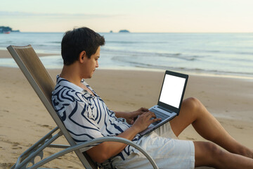 Lifestyle of young Asian man blogger working and using keyboard on laptop while sitting on the beautiful beach, freelance travel on holiday summer during sunset. Summer Vacation Concept.