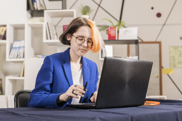Pretty young girl in blue business suit sits at table and works on laptop.