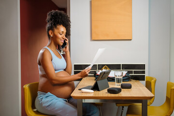 Pregnant black woman working from home at night using tablet and talking on the cellphone.
