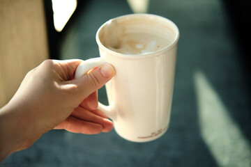 Hand holds cup of coffee, sunlight from window. Woman takes break from remote work to rest and drink coffee.