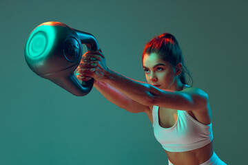 Portrait of sportive woman workout, doing exercises with sports equipment isolated on green studio background in neon light. Sport, action, fitness, youth concept. - obrazy, fototapety, plakaty