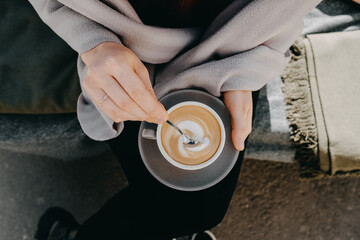 Closeup of female hands holding a cup of cappuccino, stirring foam with a spoon, top view.