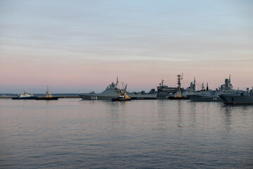 Fototapeta na wymiar Two naval harbor tugs against the background of warships in the Petrovskaya pier of Kronstadt. Evening sunset. Russia, Kronstadt, July 26, 2020