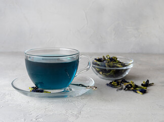 Obraz na płótnie Canvas Anchan blue tea in a transparent cup on a light table. Dry clitoria flowers in a glass bowl in the background. Light background, space for text