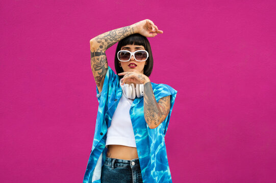 Joven mujer cauc&aacute;sica con cuerpo tatuado y  unos auriculares en el cuello sobre un fondo rosa