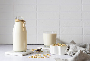 Barley milk in a glass bottle on a marble light stand. Barley grains in a white vase and milk in a glass in the background. White brick background