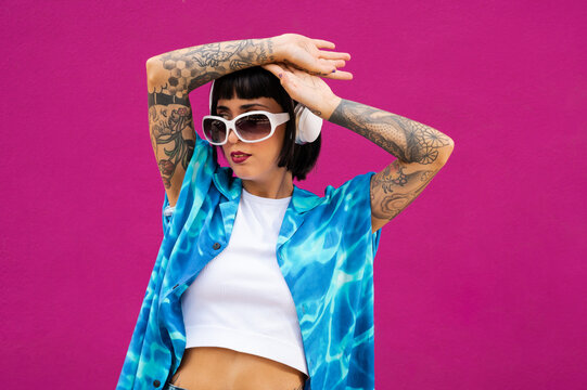 Joven mujer caucásica con cuerpo tatuado escuchando música con un auriculares sobre un fondo rosa