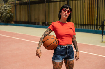 Una mujer caucásica con pelo corto y tatuajes en el cuerpo con un balón de baloncesto	 en una pista deportiva