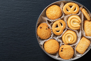 Set of crispy shortbread biscuits in an open tin over black slate surface. Tasty danish butter cookies in can for breakfast. Baked pastry, sweet food and calories concepts. Copy space.