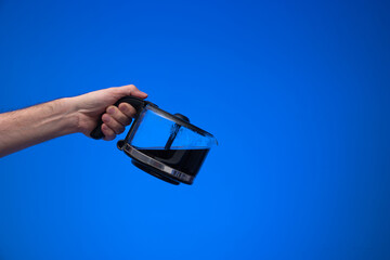 Fototapeta na wymiar Homemade large glass coffee pot held in hand by man making a pouring gesture. Close up studio shot, isolated on blue background
