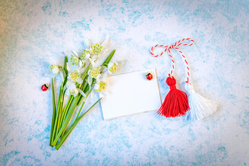 Bouquet of delicate snowdrops with Martisor talisman and blank postcard on blue background. First of march celebration Baba Marta Day. Space for text. Soft focus. Top view. - obrazy, fototapety, plakaty