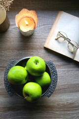 Bowl of green apples, open book, reading glasses, lit candle and flowers on the table. Top view.