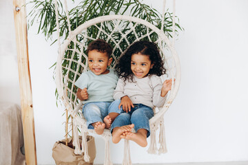 dark-skinned mulattoes A boy and a girl sit in a wicker hanging chair and ride. Children love each other and hug. - obrazy, fototapety, plakaty
