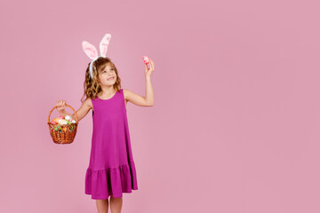 Obraz na płótnie Canvas Adorable little girl with blond curly hair in stylish dress and bunny ears headband, smiling and looking away while demonstrating wicker basket and colorful Easter egg, vertical orientation