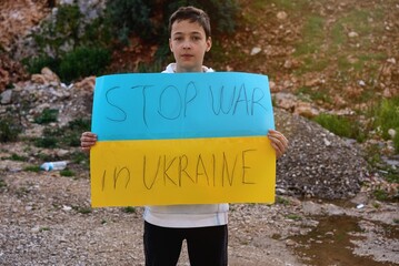 Portrait of Ukrainian boy child near the destroyed building showing banner with massage text Stop...