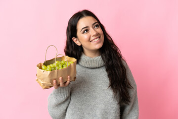 Young caucasian woman holding a grapes isolated on blue background looking up while smiling