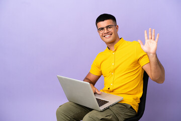 Young man sitting on a chair with laptop saluting with hand with happy expression