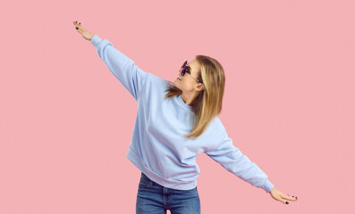 Enjoying music and catching the good vibes. Happy young woman having fun in a fashion studio. Carefree girl in sunglasses and pale blue sweatshirt dancing isolated on a pastel pink background