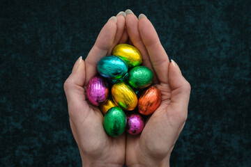 Female hands holding out a handful of small chocolate easter eggs wrapped in colorful foil. Spring holiday greeting card. Festive religious tradition. Dark green background. Pride flag colors.