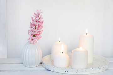 Fototapeta na wymiar A bouquet of pink cut hyacinth a in a small white corrugated vase and three large burning candles on a round tray are on a beige table. Place for text