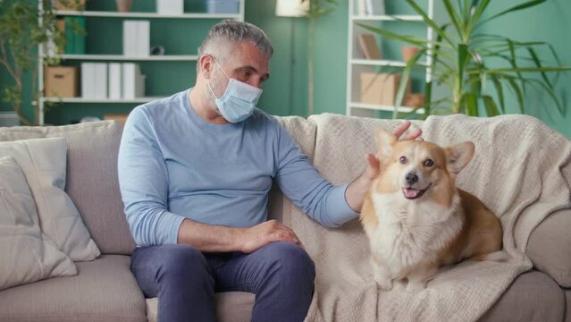 A Sad Lonely Elderly Man in a Medical Mask Stroking His Dog While Sitting on the Couch at Home. Canistherapy, Best Friend, Help, Pet, Mental Health. Pandemic, Social Distancing and Self-isolation.