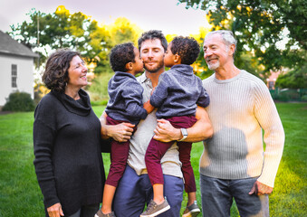 Portrait of three generations of of mixed race family in Midwestern park; white grandparents, father and African American twin boys; boys kissing their father; grandparents look at them 