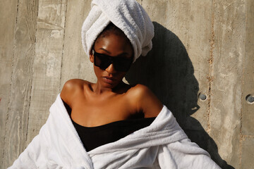 Pretty young black woman wrapped in white towel posing over grey wall. Outdoor.