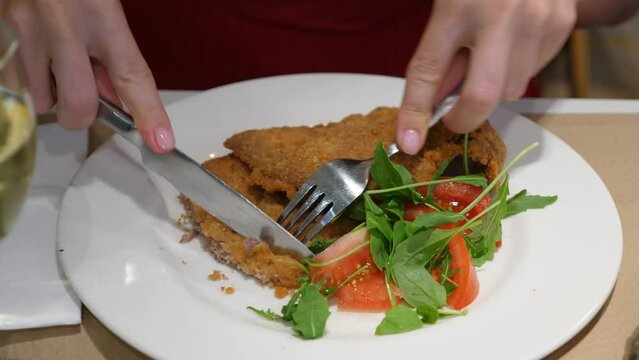 Woman using knife and fork to cut little piece from breaded beef escalope, closeup shot. Meal served with green Leaves of Arugula and tomato slices. Popular Milanesa meal served as main or second dish