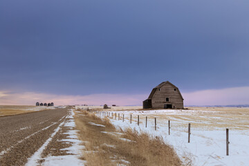 Prairie winter farm road leading into distant grain silos and dark sky. An old barn sits in farm...