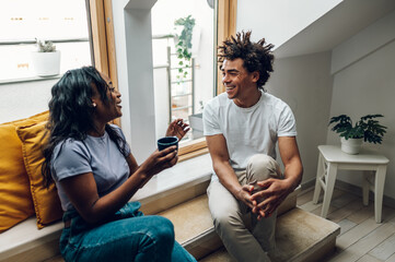 African american couple sitting on the windowsill and drinking coffee at home