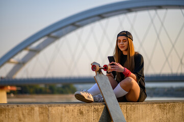 Young woman sitting near the river and using smartphone after longboard ride